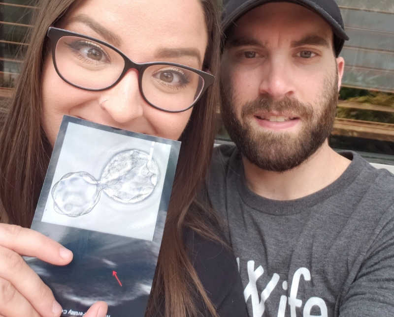 Wife holds up picture of implanted embryos in front of her face beside husband in selfie