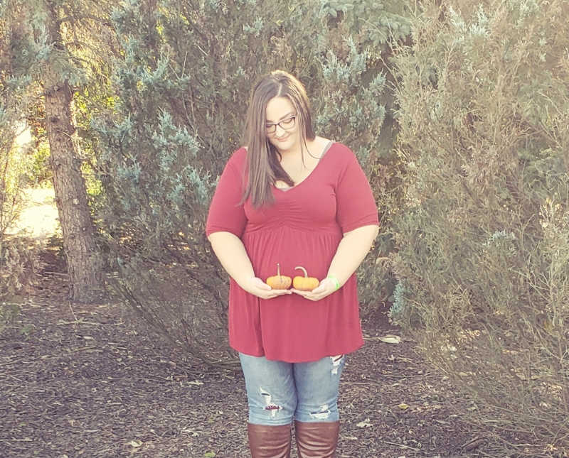 Pregnant woman stands outside holding two little pumpkins in front of her stomach