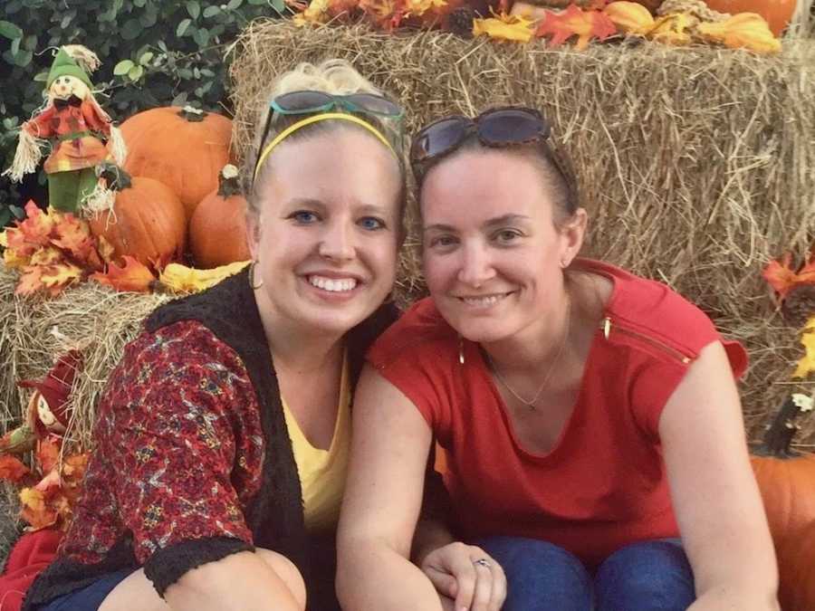 Lesbian couple sits smiling on ground in front of hay bales