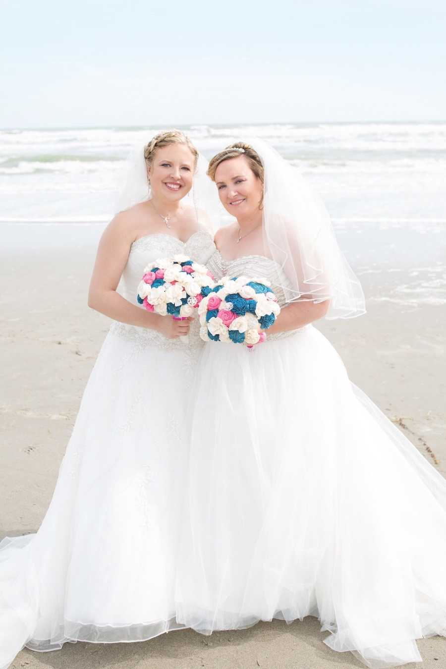 Lesbian brides stand on beach holding bouquet of colorful flowers