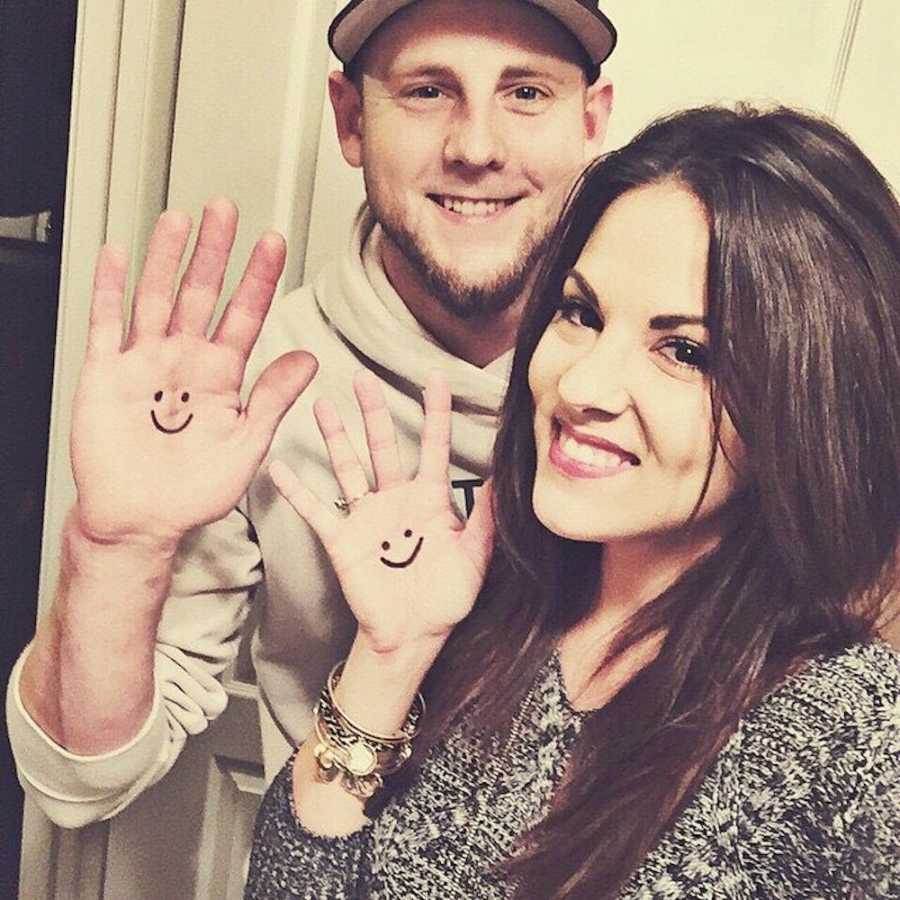 Husband and wife who want to adopt stand smiling holding up their hands that has smiley faces drawn on them