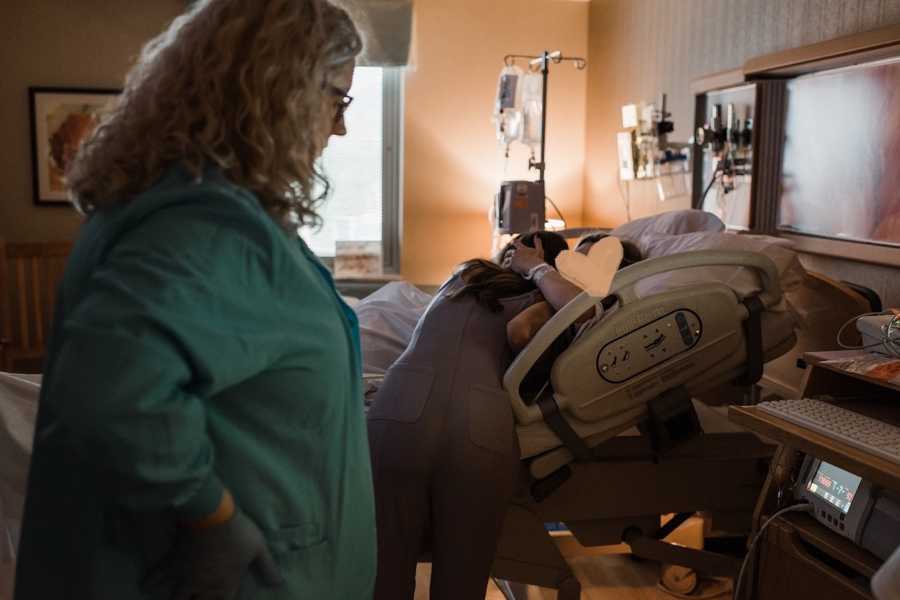 Nurse stands at machine in hospital room while woman hugs mother of her child in hospital bed