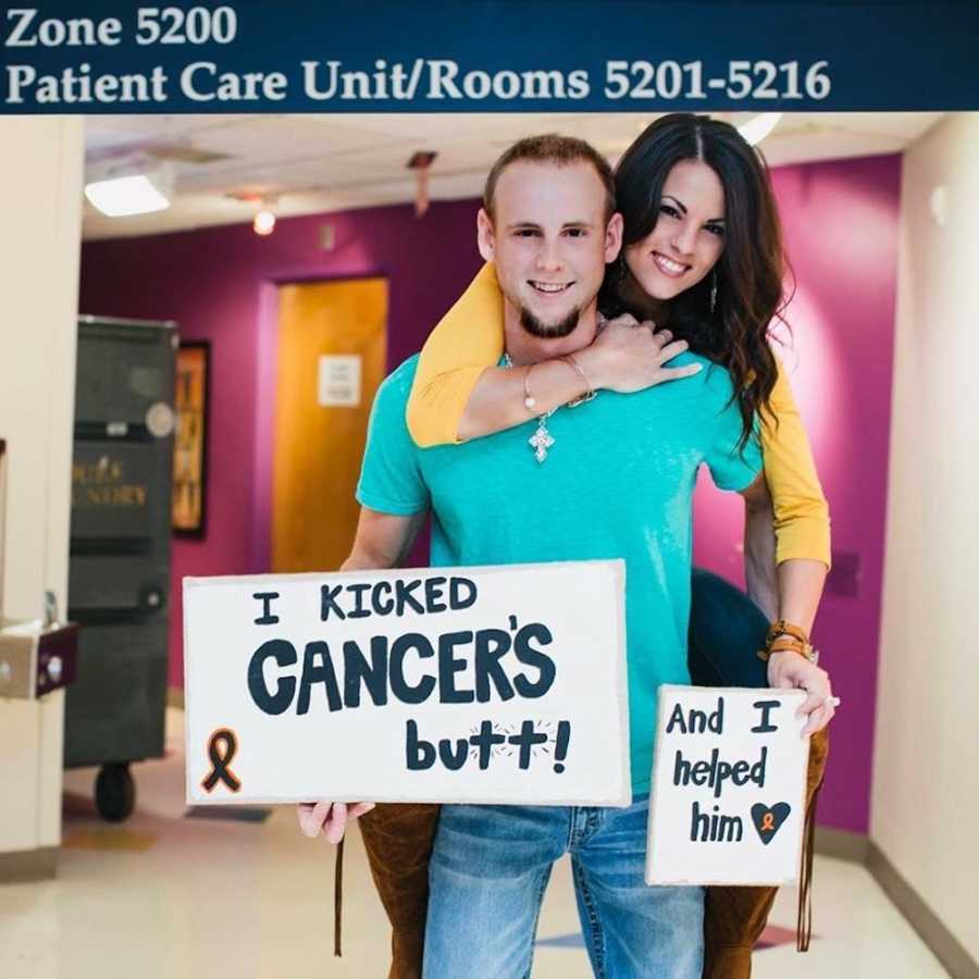 Young man who beat cancer stands in hospital hallway holding sign while fiancee smiles on his back