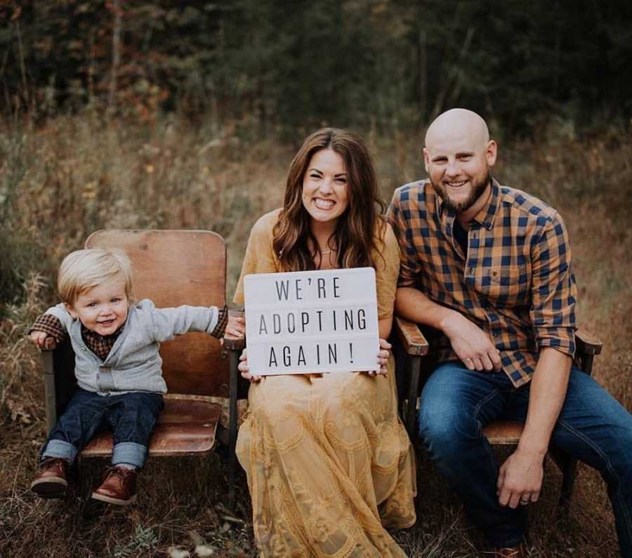 Husband and wife sit beside their son outside in stadium chairs holding sign to announce second adoption