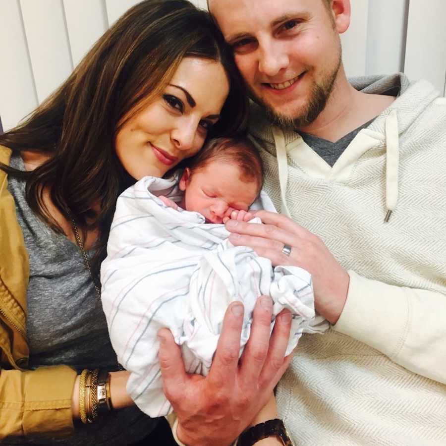 Husband and wife smiles as they hold swaddled newborn in their arms