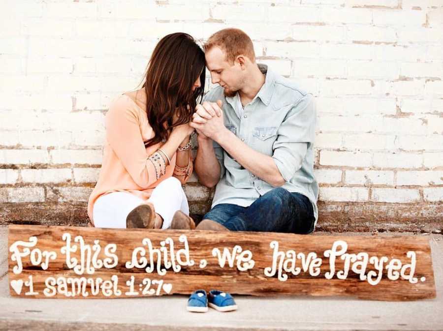 Husband and wife sit on ground leaning against white brick wall holding hands as they pray for child