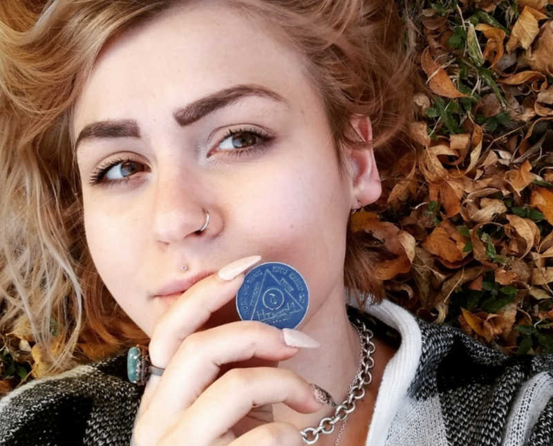Woman lays on ground outside holding up her sober chip in selfie
