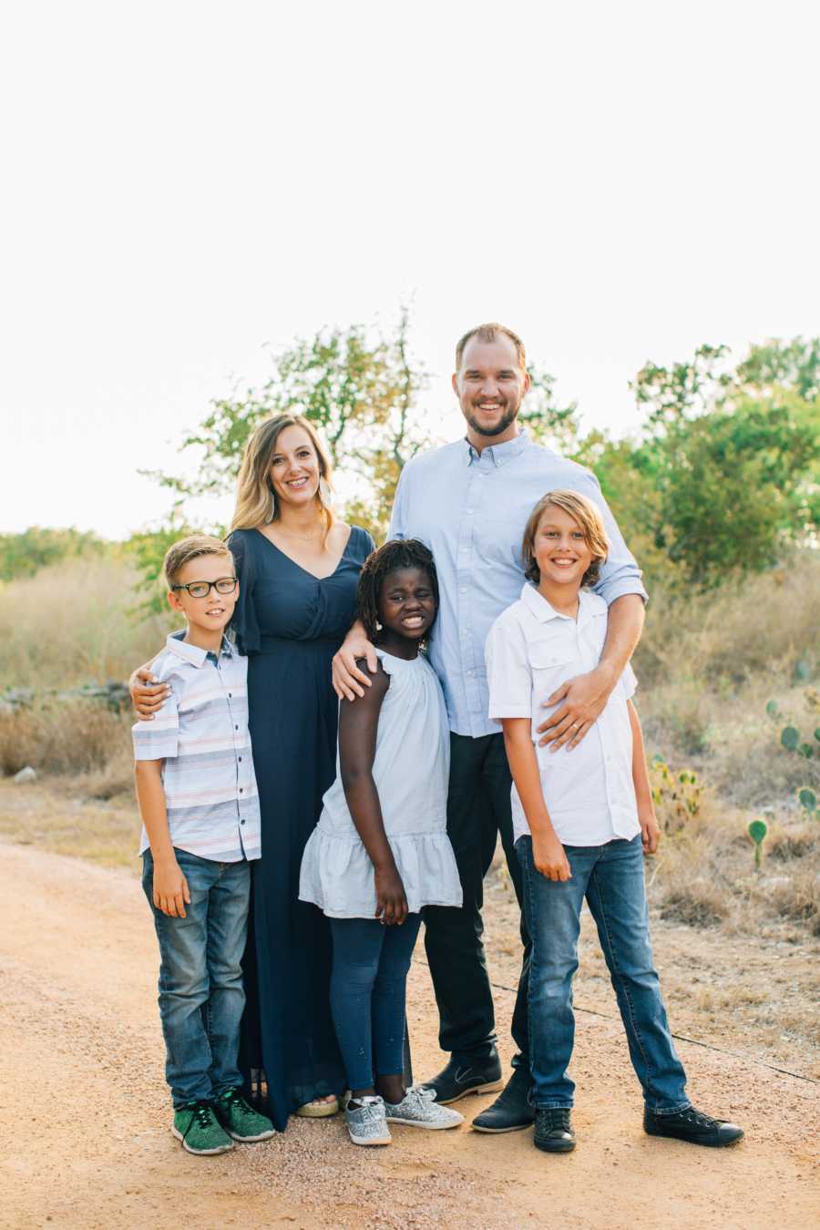 Husband and wife stand on dirt path outside with two sons and adopted daughter
