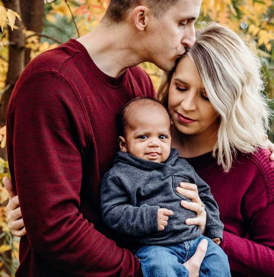 Husband holds adopted baby outside as he kisses wife on forehead in photoshoot