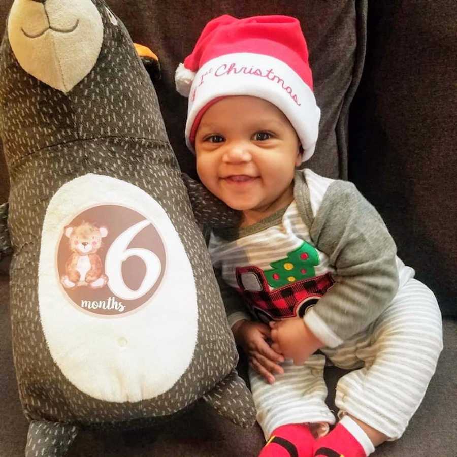 Adopted baby wearing Santa hat sits on couch in Christmas pj's beside stuffed animal that says, "6 months"