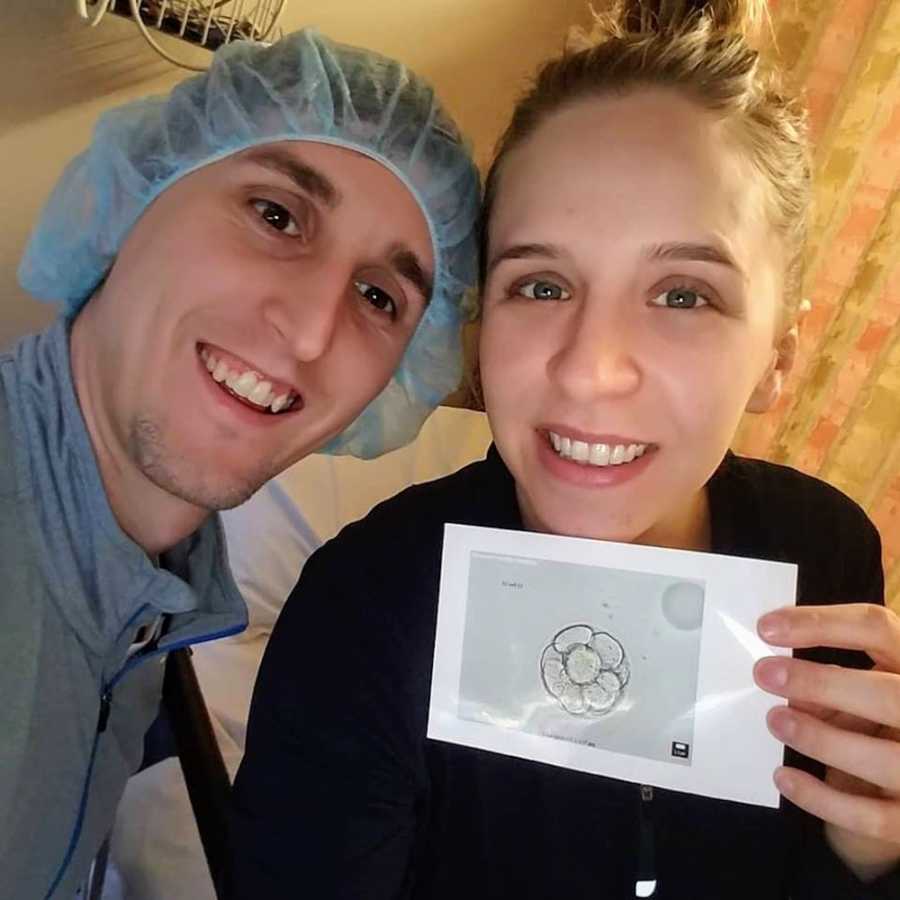 Husband wearing hairnet smiles beside wife who holds ultrasound of fertilized egg