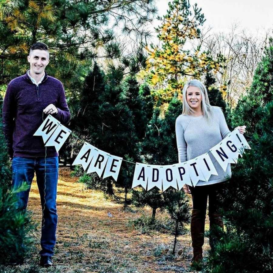 Husband and wife who struggled to get pregnant stand outside holding sign that says, "We are adopting"