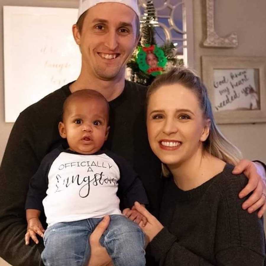 Husband smiles holding adopted baby as he has arm around smiling wife in home