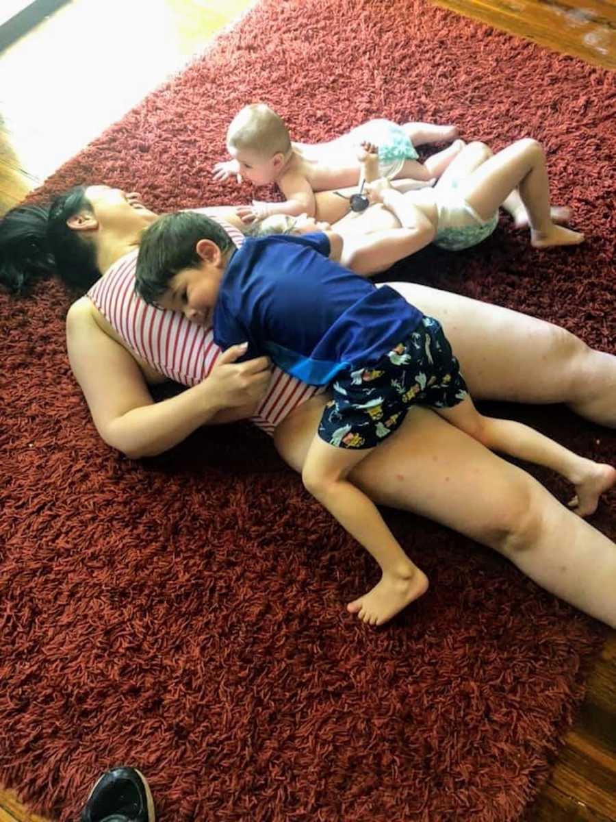 Woman lays on carpet with toddler son laying on her and baby beside her