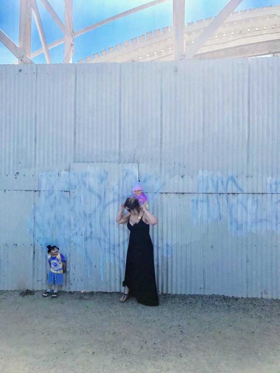 Mother stands outside with daughter on her shoulder and son standing beside her