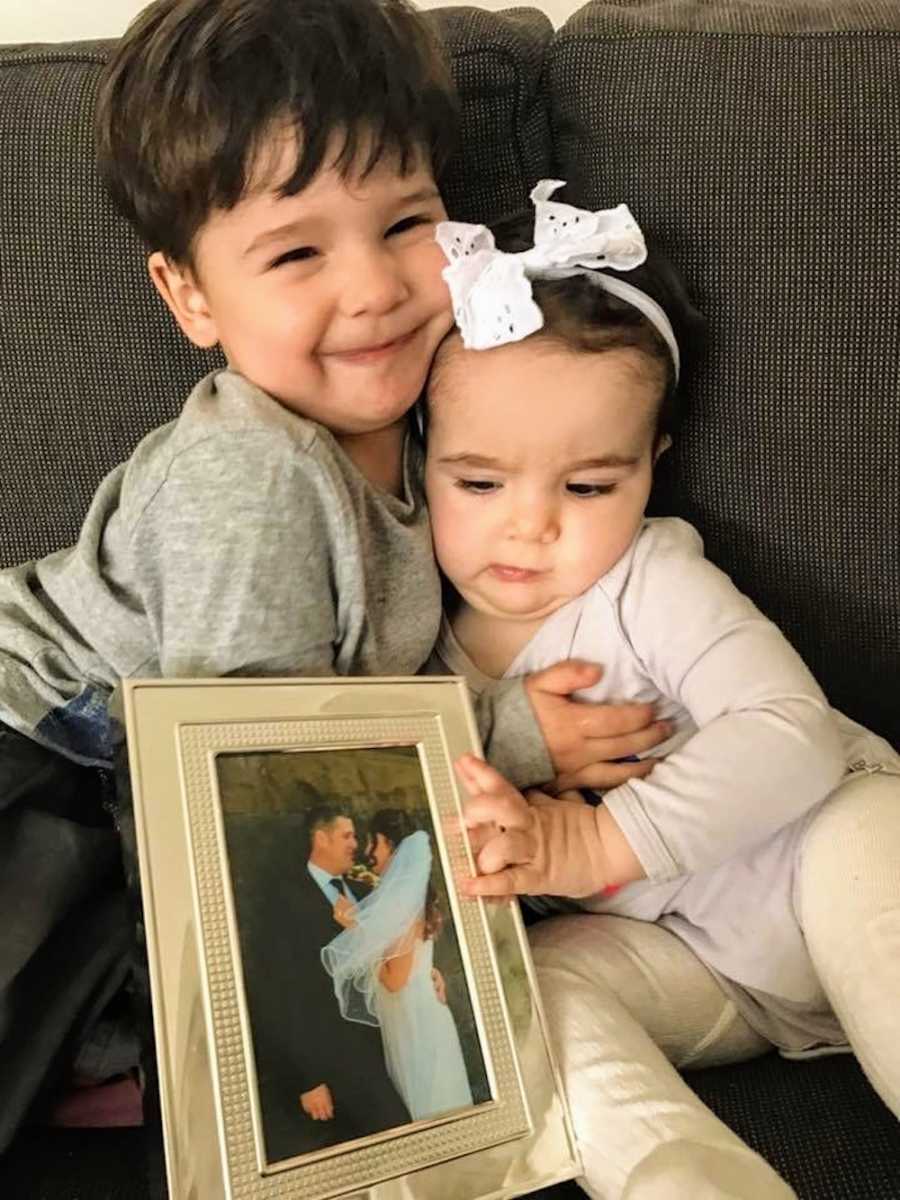 Little boy sits on couch hugging baby sister who holds picture frame of parents on wedding day