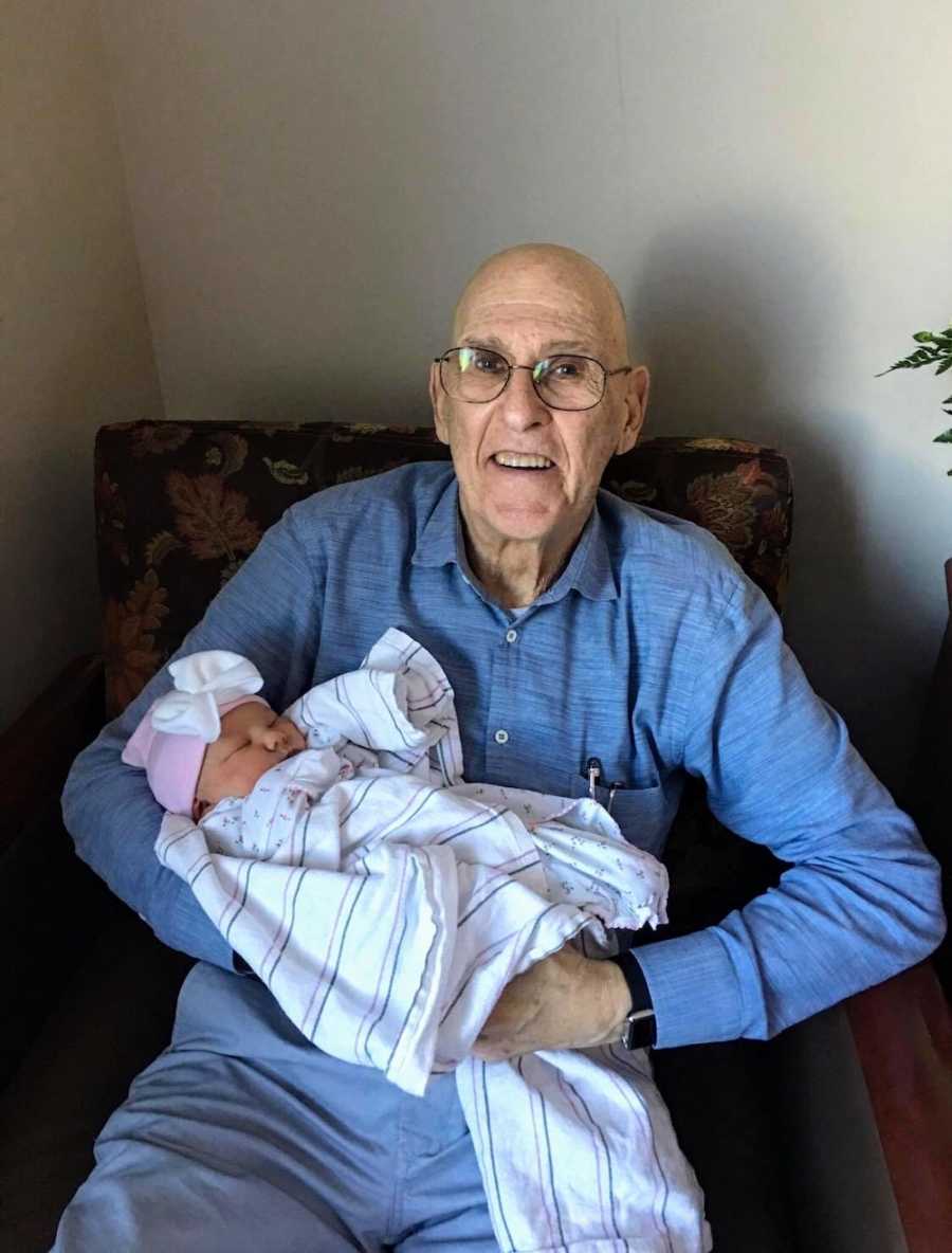 Elderly man sits in chair smiling while he holds baby girl wrapped in blanket