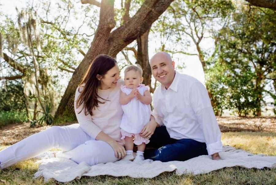 Husband and wife sit on blanket outside with their baby daughter standing between them 