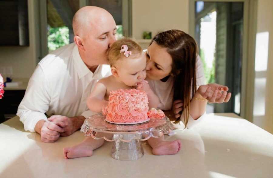 Baby girl sits on kitchen counter eating cake while her parents stand behind her kissing her head