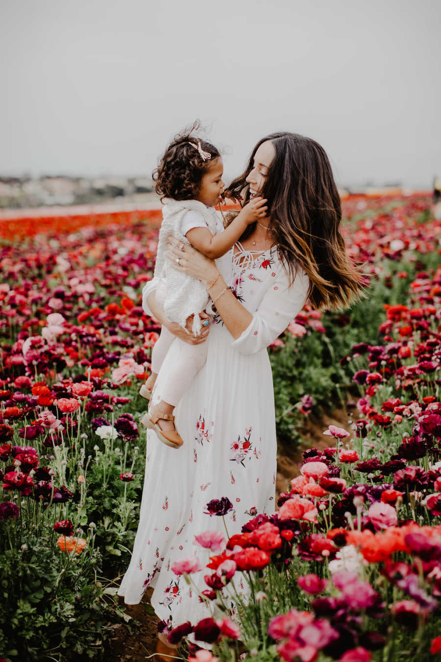 Mother stands in field of pink and red flowers holding adopted daughter