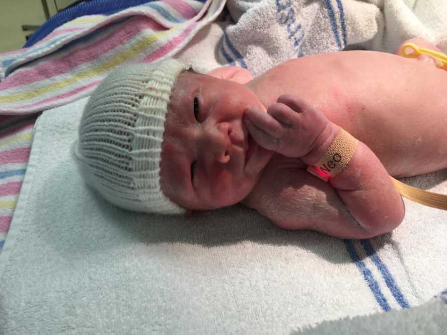 Newborn with white hat on lays on back in hospital holding hand to mouth