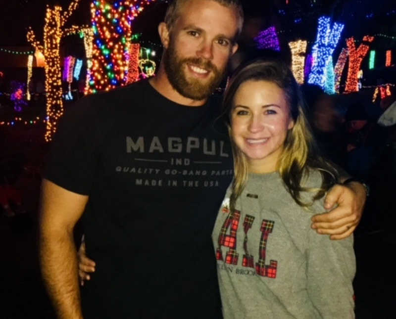 Husband and wife smile as they stand arm in arm outside in front of trees decorated with Christmas lights