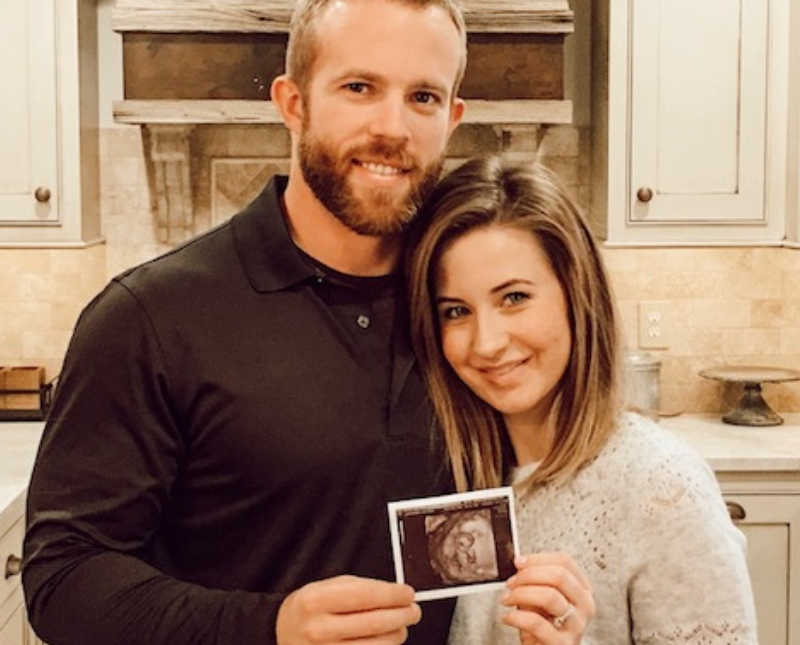 Husband and wife stand smiling in kitchen as they hold ultrasound picture of their baby