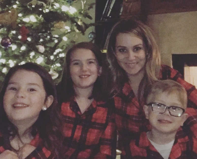 Mother smiles with three children in matching pj's in front of Christmas tree