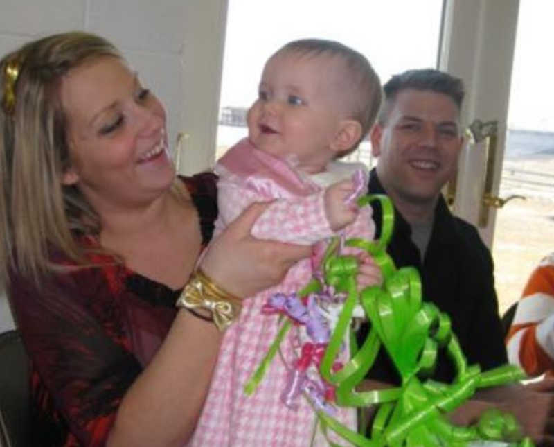 Mother sits at table smiling at baby daughter standing in her lap while she sits beside husband