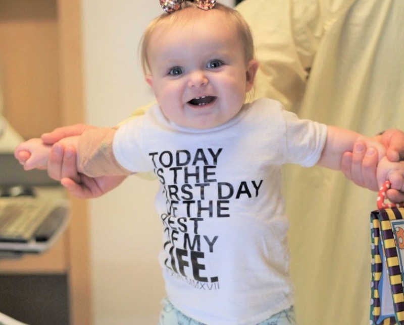 Little girl with Bubble Baby Disease stands smiling in arms of parent