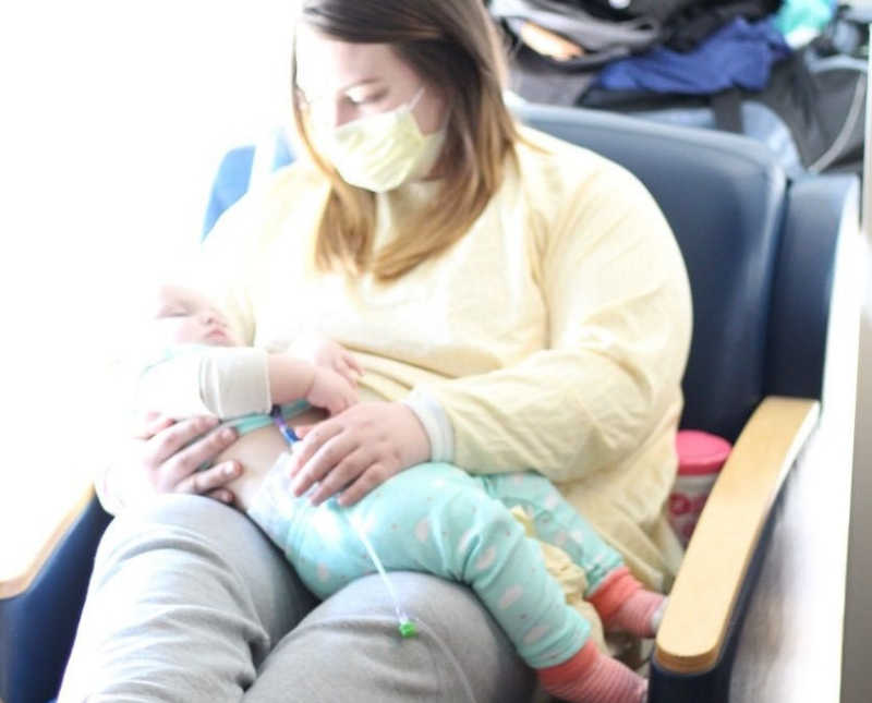 Mother sits in hospital with mask on holding daughter in her lap who has Bubble Baby Disease