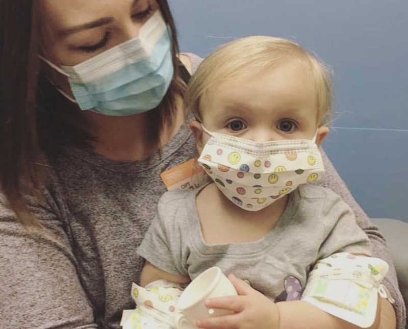 Mother with mask on looks at daughter with Bubble Baby Disease who sits on her lap