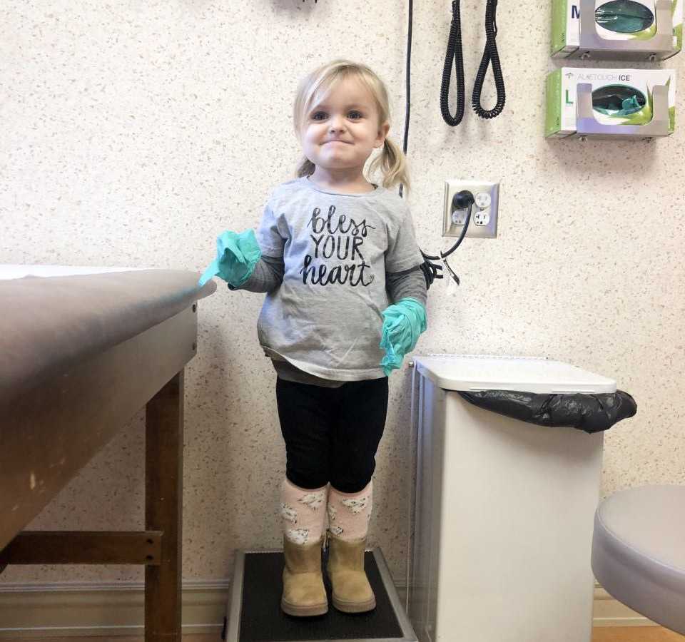 Little girl who had Bubble Baby Disease stands on stool in doctor's office smiling