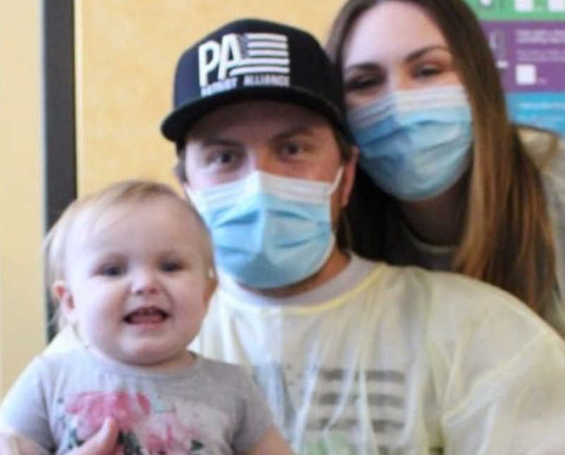 Wife with mask on stands behind husband with mask on who has daughter with Bubble Baby Disease on his lap
