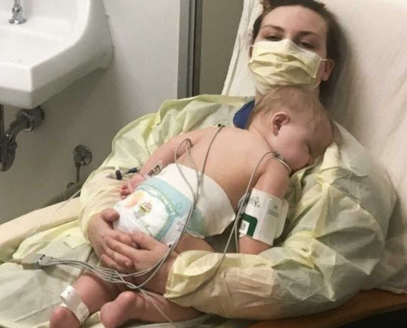 Mother lays in hospital bed with mask on holding baby with Bubble Baby Disease