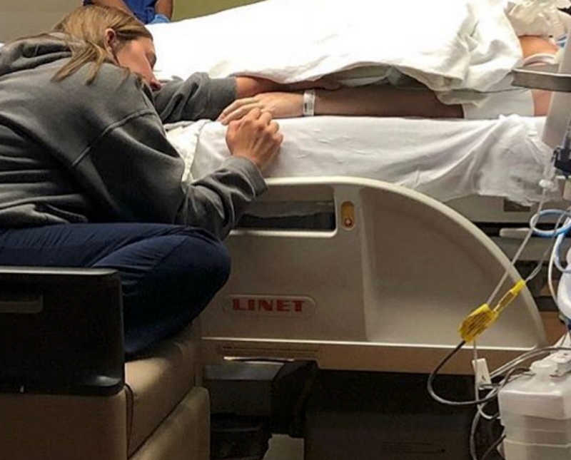 Woman sits in chair in hospital beside bed where husband with tear in aorta lies