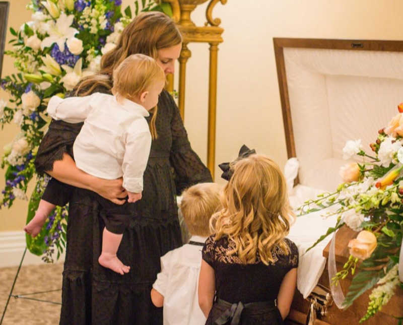 Woman stands holding son beside two young kids at casket of their faither
