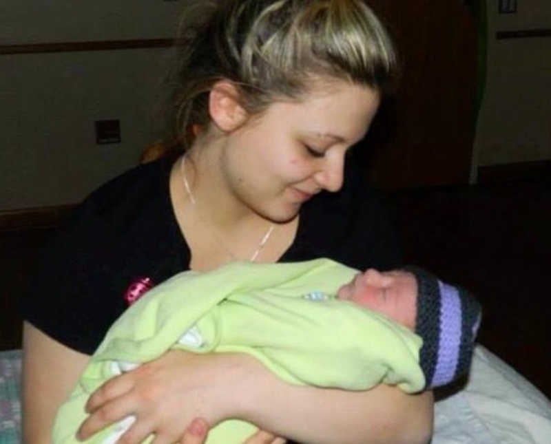 Teen mother smiles as she looks newborn asleep in her arms
