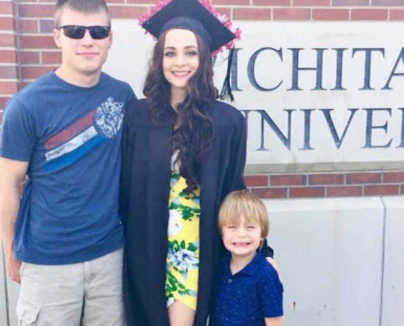 College grad stands smiling in cap and gown beside boyfriend and her young son