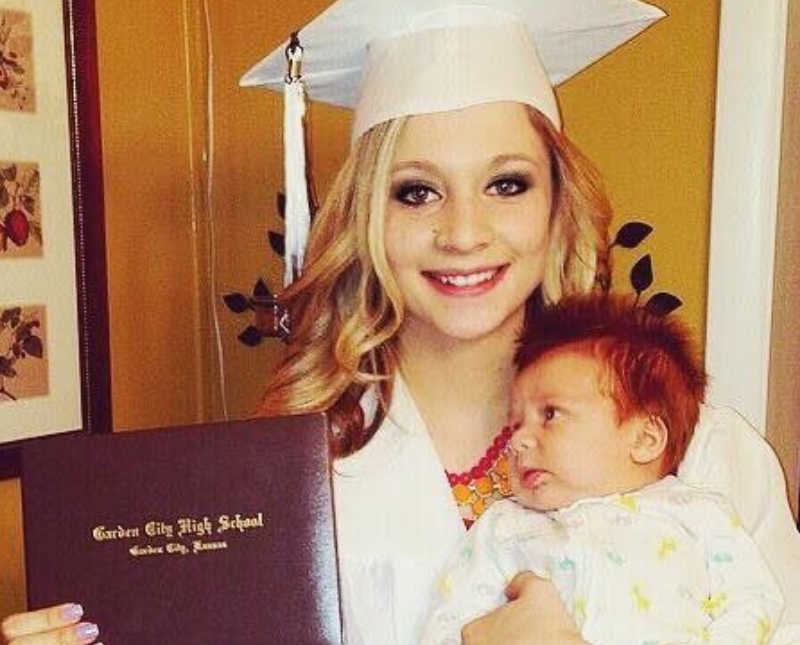 High school graduate stands in cap and gown holding her baby on one arm and diploma in other