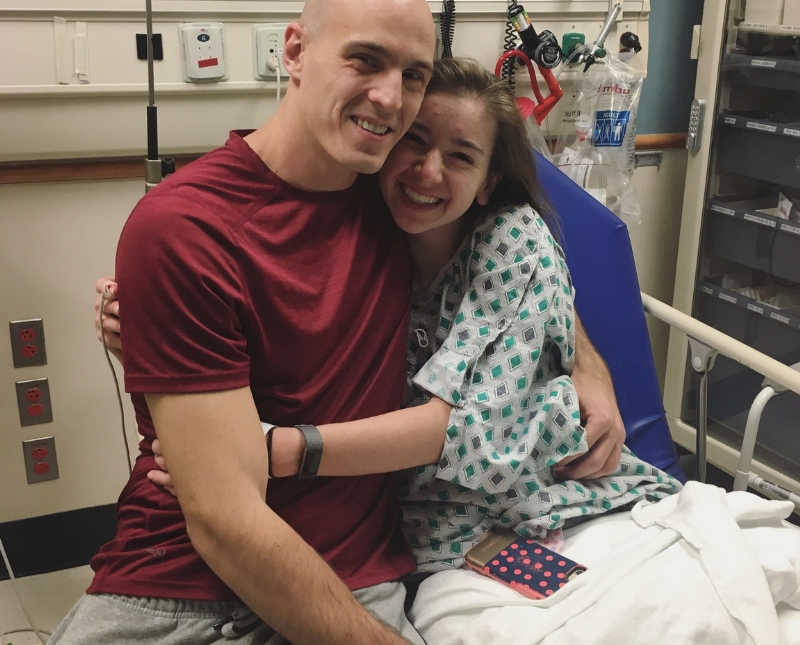 Woman with chronic illness sits in hospital bed hugging man 