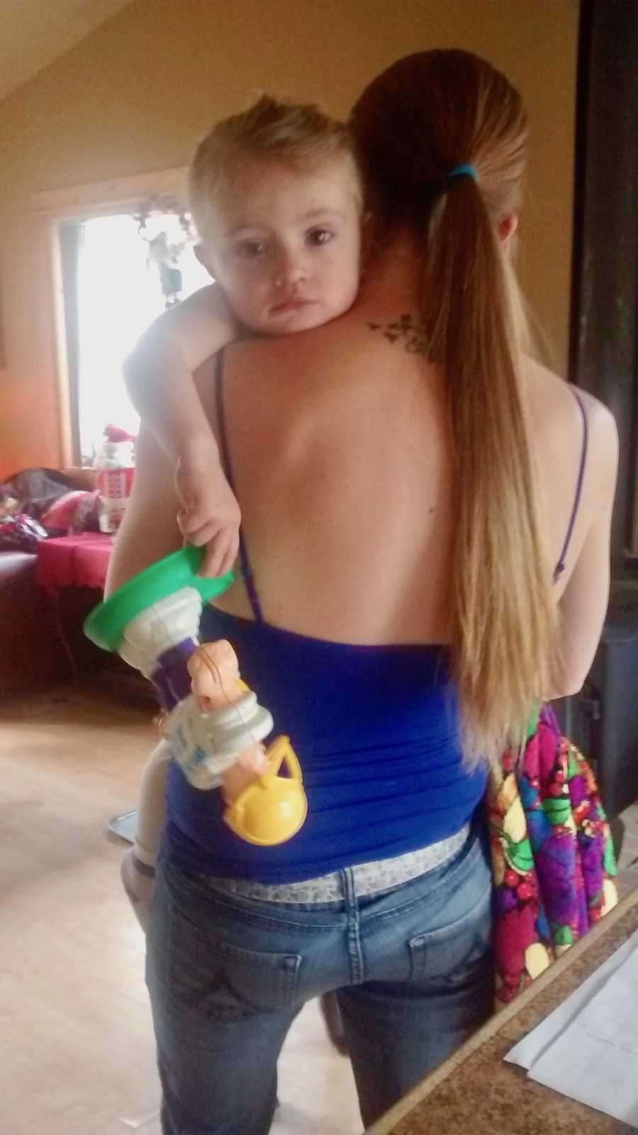 Mother stands holding toddler daughter while she holds a toy 
