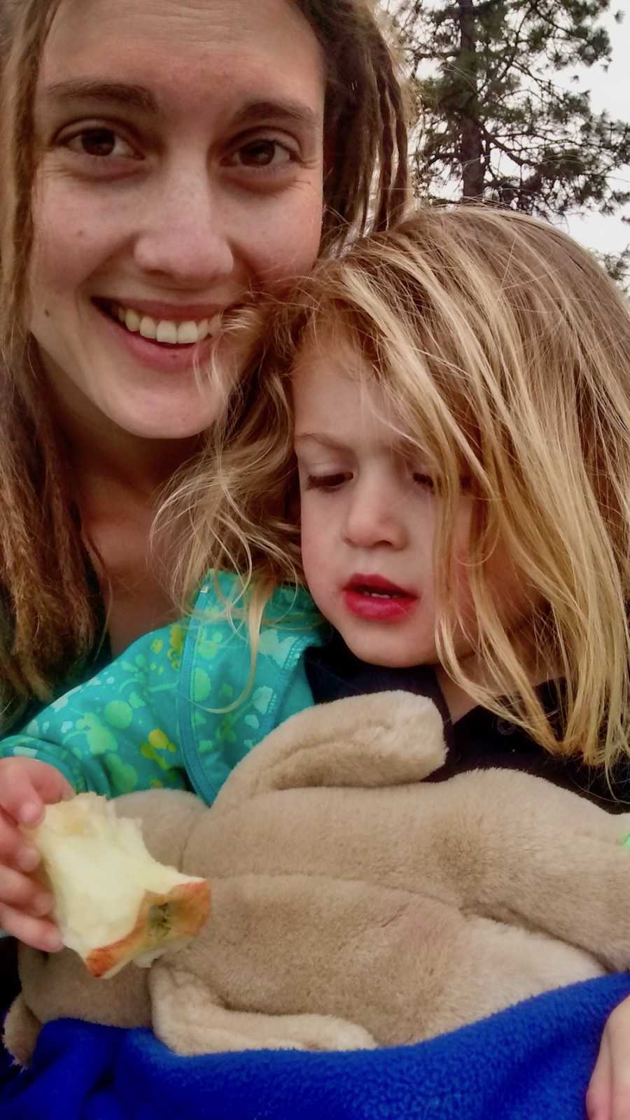 Mother smiles in selfie with her daughter in her lap who holds onto apple