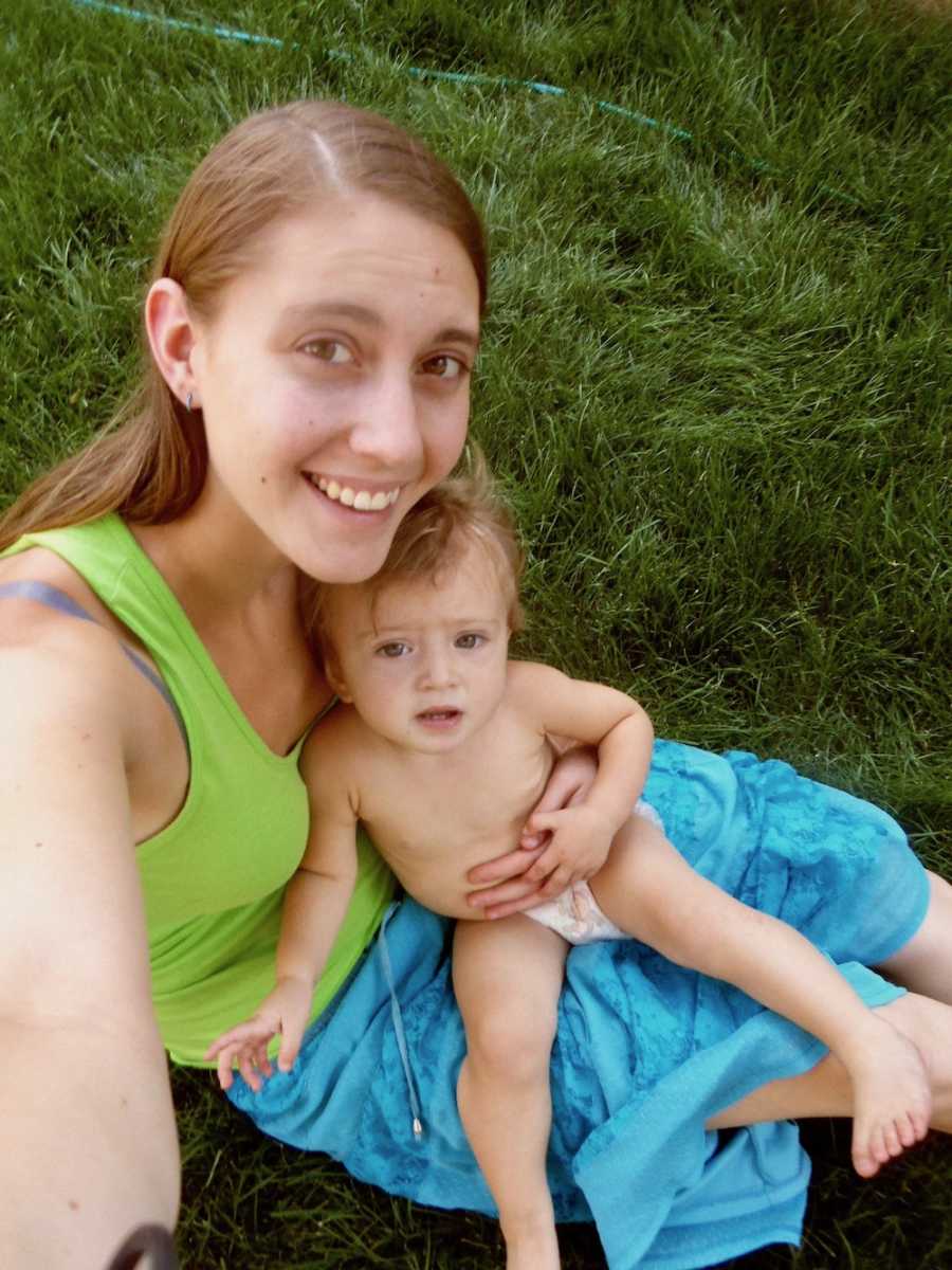 Mother smiles as she sits on ground outside with daughter in her lap