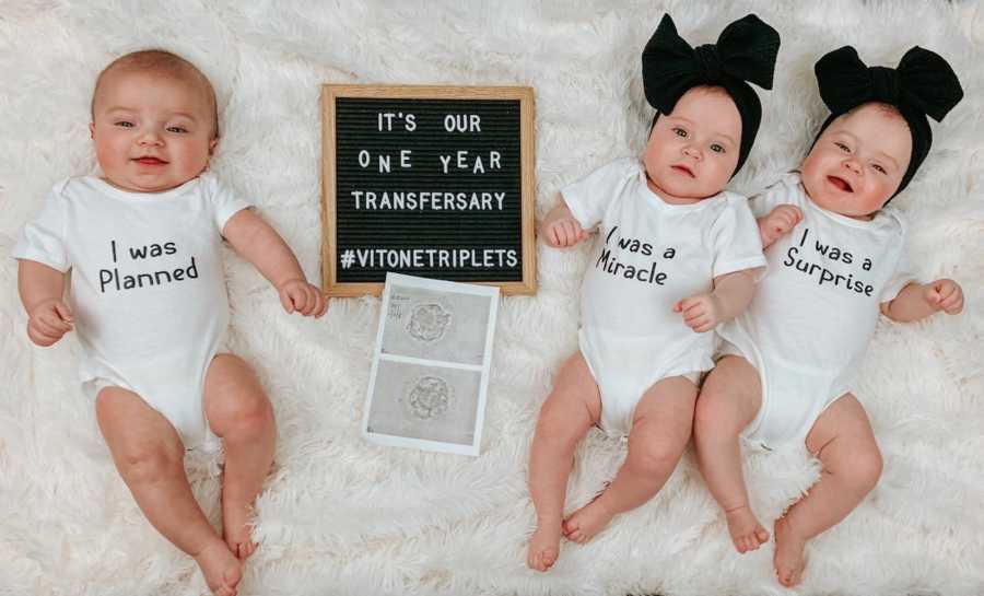 Triplets lay on back on white rug with sign beside them that says its been one year since their mom had embryo transfer