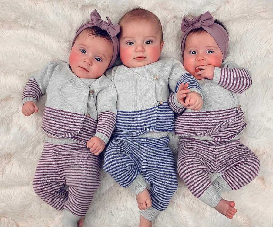 Two girl triplets lay in purple and gray outfit with their brother between them in blue and and gray outfit