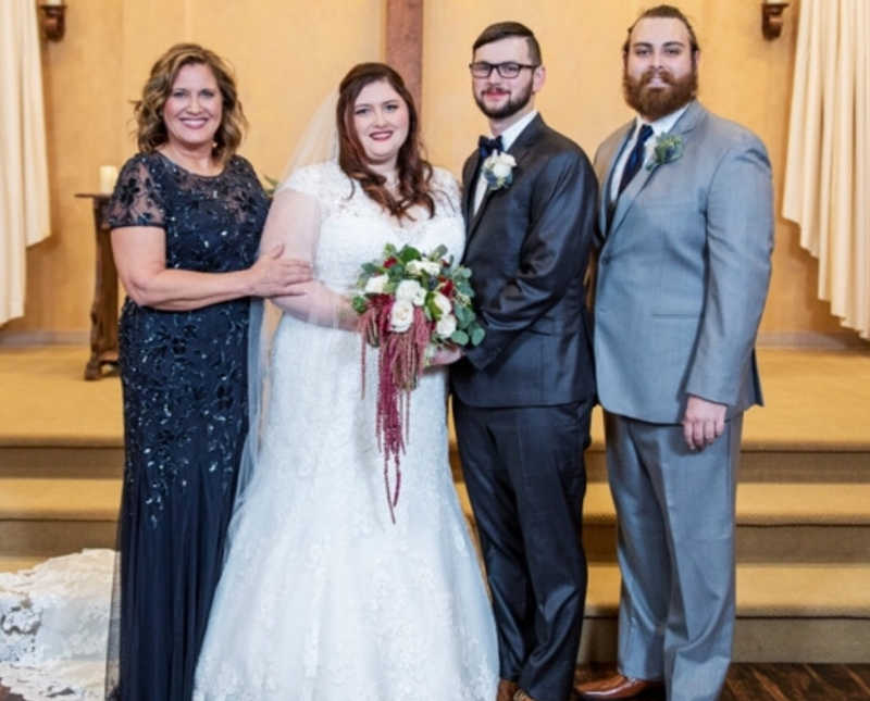 Bride stands at altar smiling with groom, mother, and brother