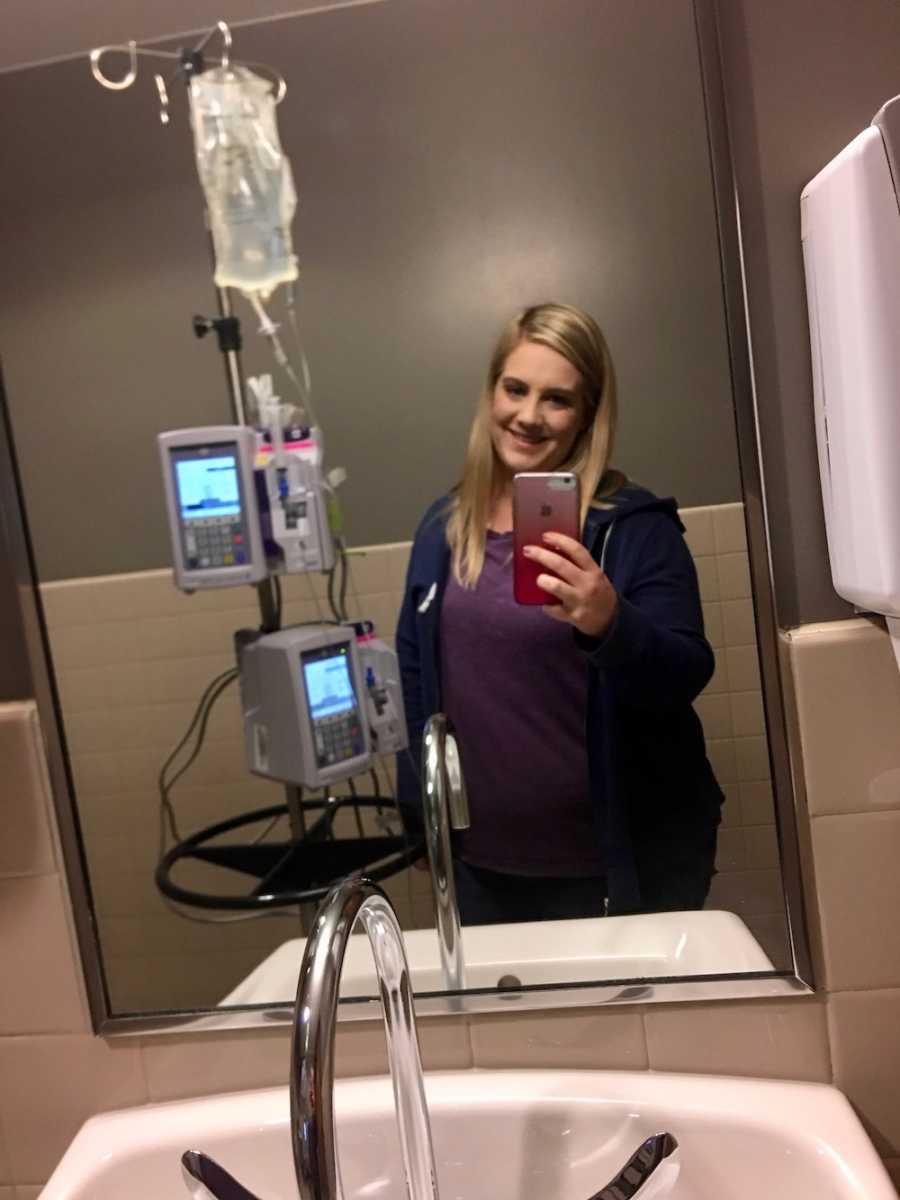 Woman with Crohn’s Disease takes mirror selfie in hospital bathroom with IV bag at side