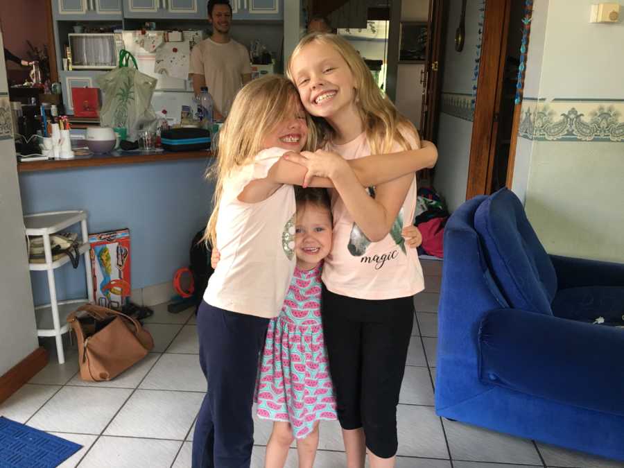 Young girl who was almost died stands in home hugging two younger sisters
