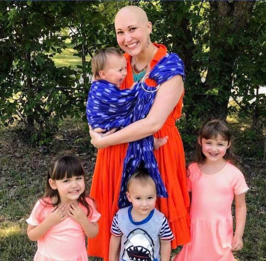 Mother with Acute B-Cell Lymphoblastic Leukemia stands outside with baby swaddled to her chest beside three other kids